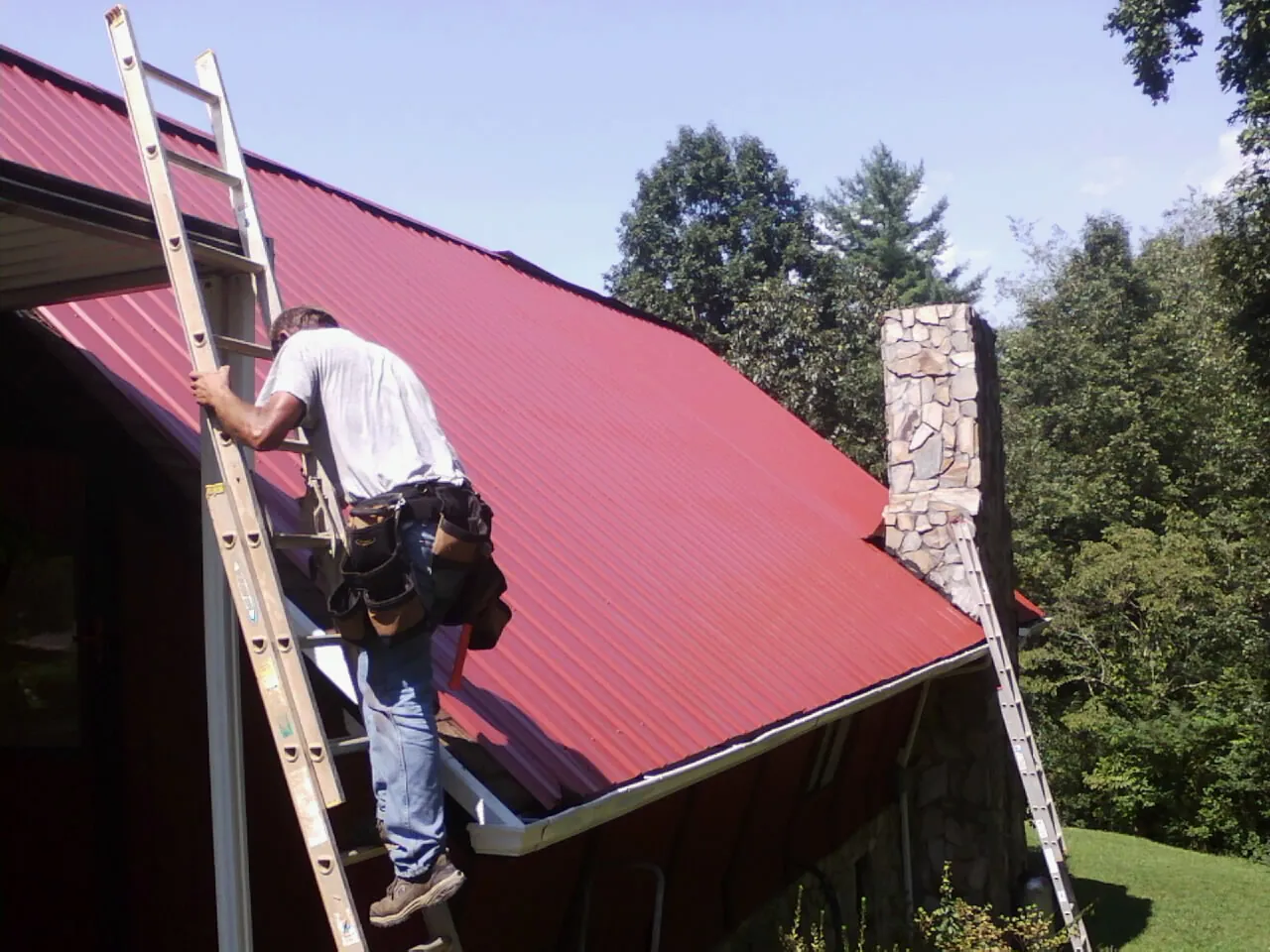 Roofer on a ladder beside a red metal roof and stone chimney — Soffit & Fascia Repair by Roofing Brothers in Summer Lake